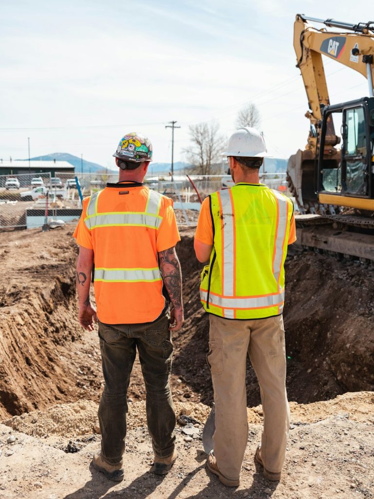 Two construction workers in high-visibility safety clothing review an in-progress excavation at a jobsite.